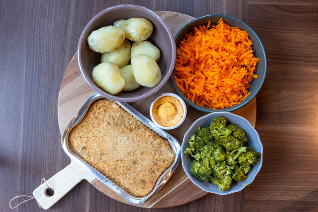 Et rundt skjærebrett med hank, oppe står en rektangulær aliminiumsform med fiskegrateng, en skål kokte poteter, en skål råkost av gulrøtter, en skål kokt brokkoli og et lite plastbeger med kryddersmør på. 