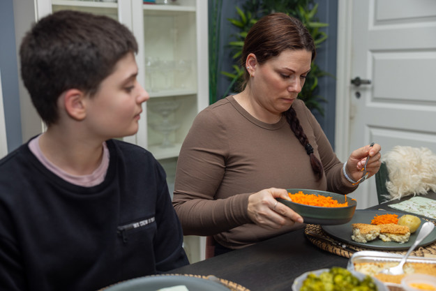 Tenåring med kort sort hår og sort genser ser mot mot en voksen kvinne med brunt hår ikledd en brun langermet topp, hun holder en skål med raspet gulrot i ene hånden og en skje hun bruker til å ta raspet gulrot med i den andre hånden. hun ser ned på gulroten. 