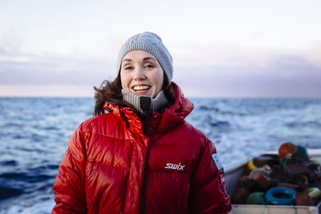 Smilende kvinne, med mørkt hår, grå lue. Hun står utendørs med havet i bakgrunn. På seg har hun en tykk rød jakke. 